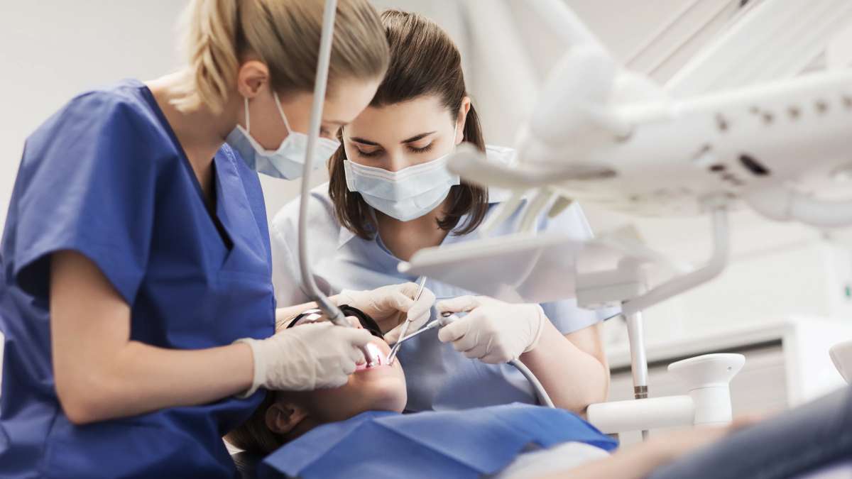 Two dental professionals wearing scrubs, gloves, and face masks performing a dental procedure on a patient lying in a dentist’s chair, with dental instruments and overhead equipment visible.