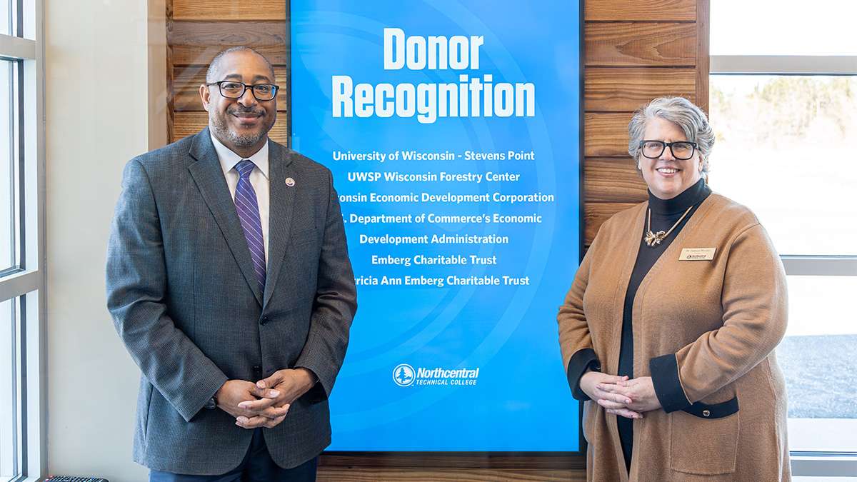 Two presidents from organizations smiling in front of a donor recognition sign at Northcentral Technical College.
