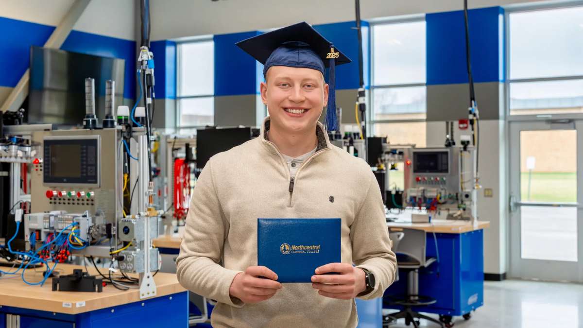 Ethan Sondelski smiling while wearing his graduation cap and holding his diploma while standing inside the Industry 4.0 Smart Manufacturing Lab