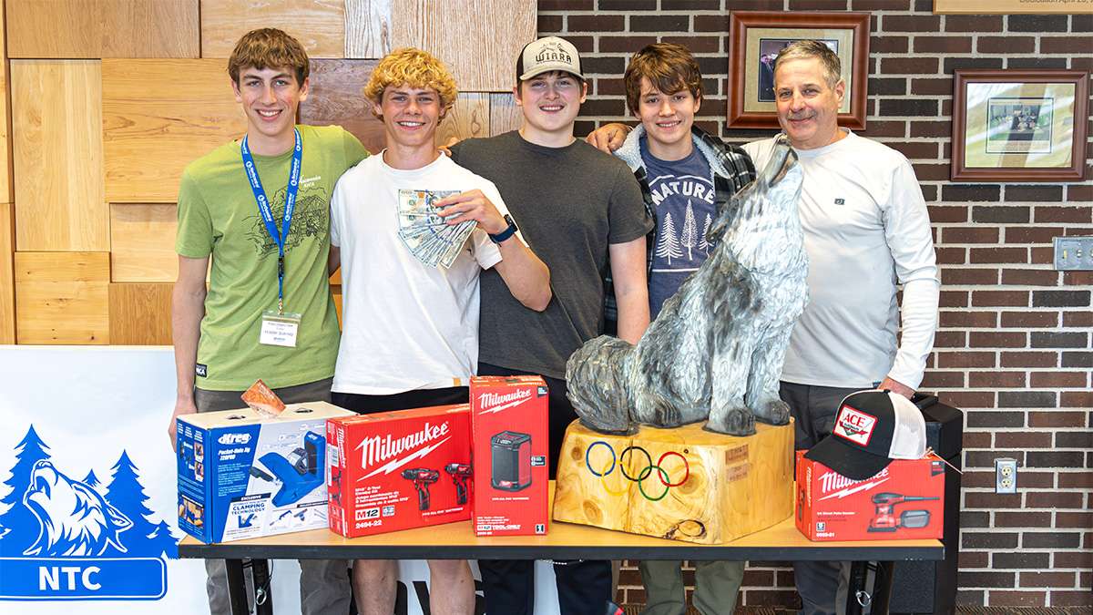 Students and instructor pose with tools and Olympic-themed wolf sculpture at technical college workshop.