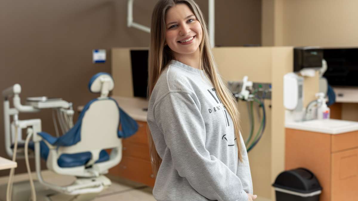 A smiling student stands in a dental lab setting, with a dental chair and equipment visible behind her, wearing a casual sweatshirt and looking toward the camera.
