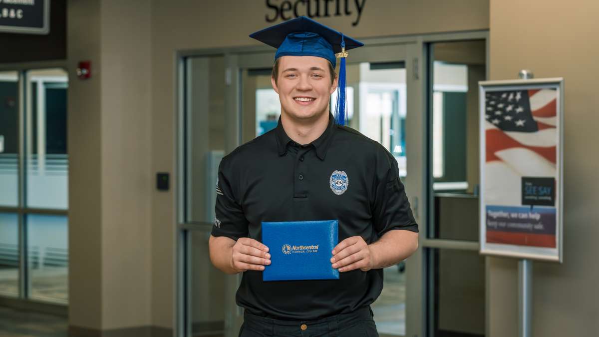 Kyle smiling while wearing his graduation cap and holding up his diploma in front of the Safety and Security Office.