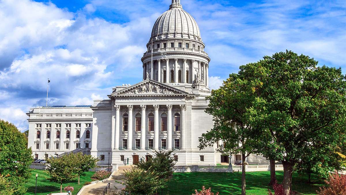 External view of the capitol building in Madison Wisconsin with grand white columns, manicured green lawn and trees.