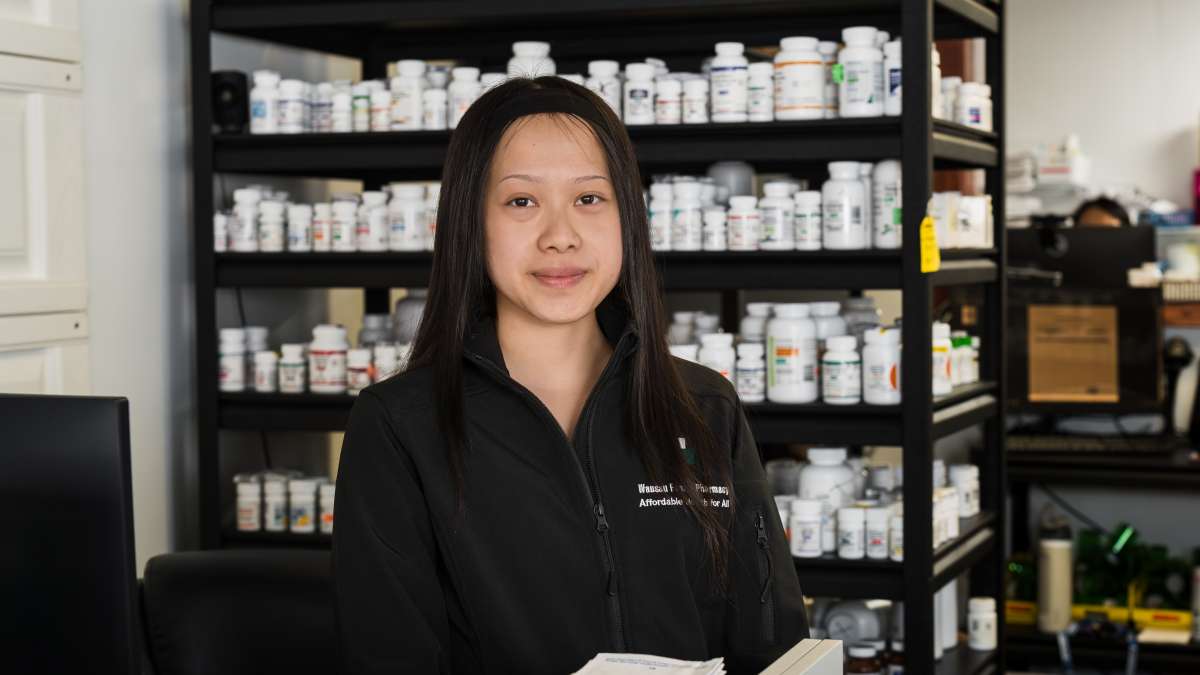 Nancy Xiong, a pharmacy technician youth apprentice, stands behind a pharmacy counter, looking toward the camera, with shelves of prescription medication bottles neatly organized behind her. She wears a black pharmacy-branded jacket and is positioned in a working pharmacy environment that includes medication storage, dispensing supplies, and computer workstations.