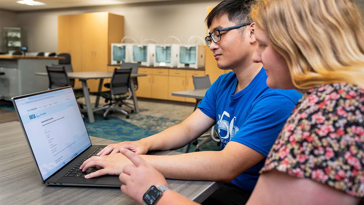 A student and faculty member collaborate at a laptop in a bright computer lab; a young man types while a young woman watches.