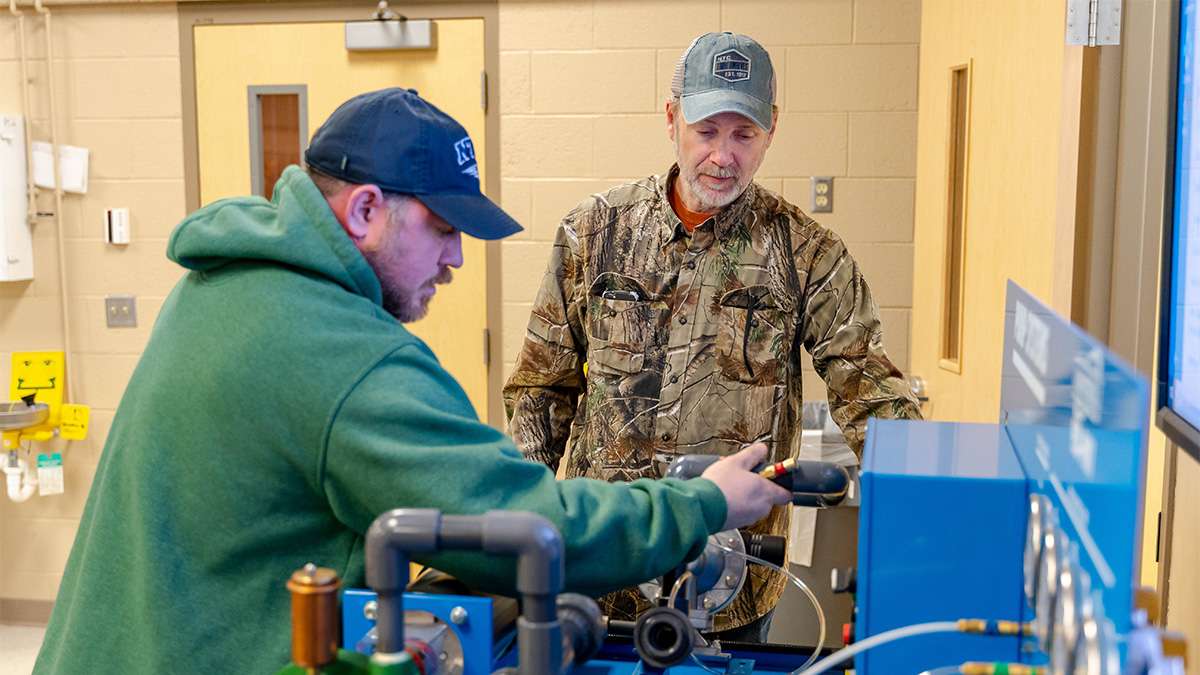 Two men focused on metal machinery in a workshop: one in a green hoodie adjusting equipment, another in camouflage jacket.