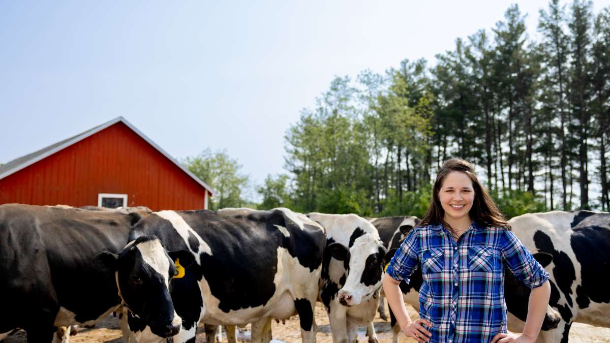 A photo shows a person standing with hands on their hips beside a group of black-and-white cows. A red barn and a line of trees are visible in the background.