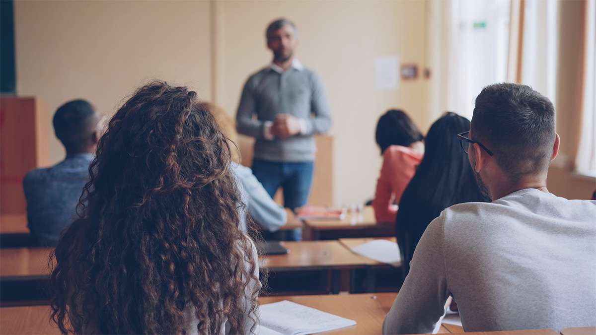 College students seated at desks facing a standing instructor at the front of a sunlit classroom.