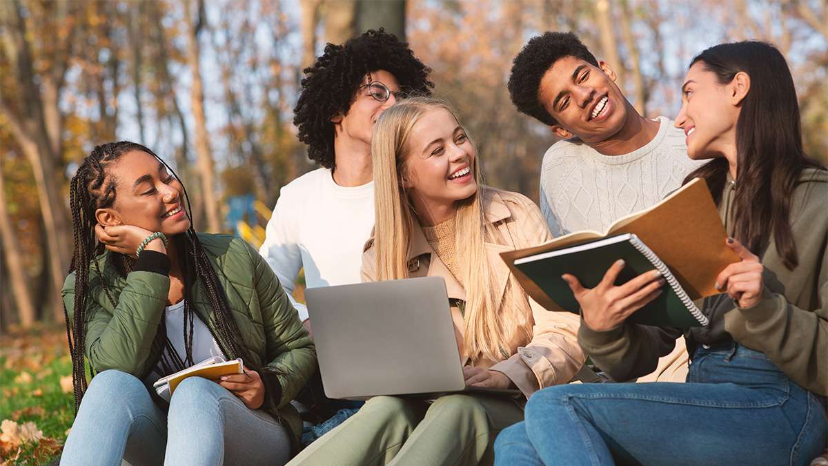 Five diverse college students sit on grass in an autumn park, laughing and studying together with notebooks and a laptop.