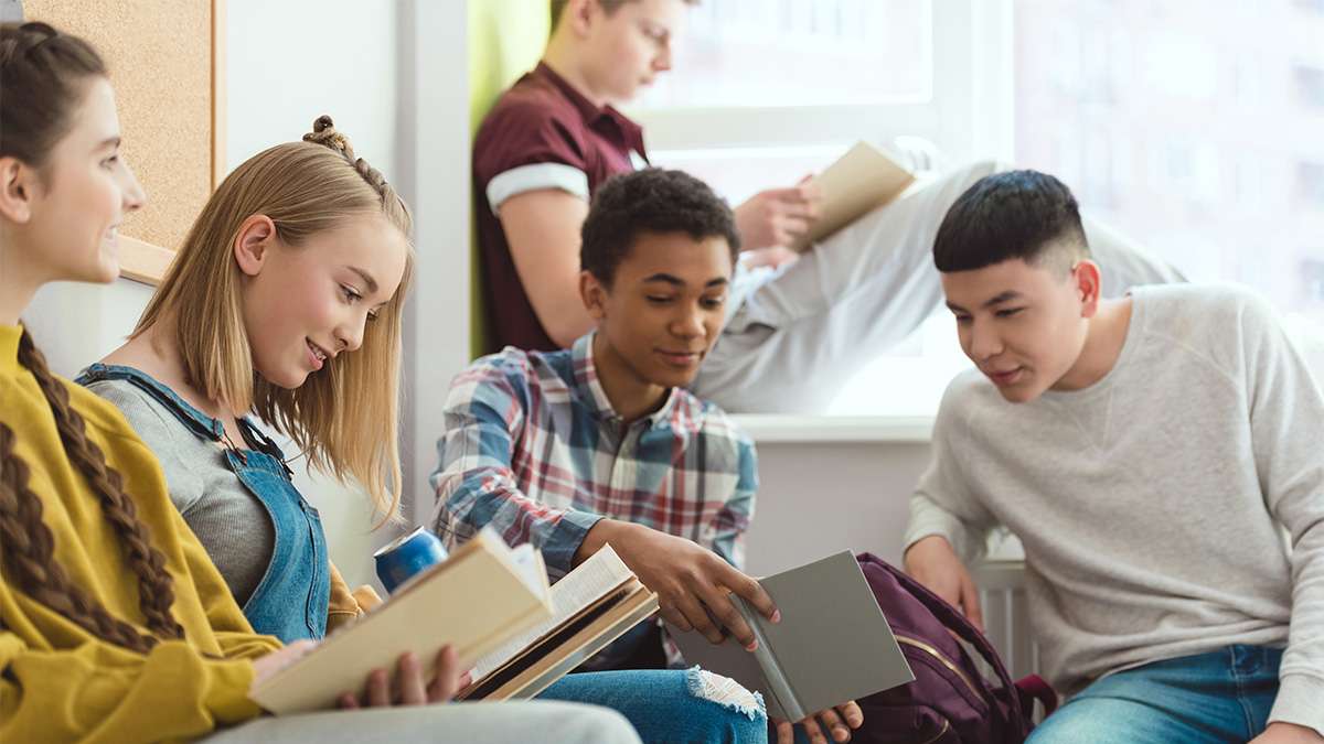 Five diverse teenagers in a bright classroom, casually seated by a window, smiling while reading open textbooks.