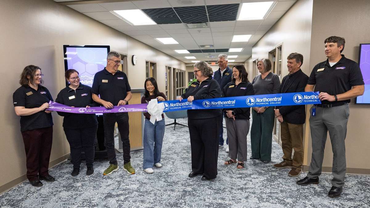 Group of people in an office hallway cutting a long blue ceremonial ribbon with oversized scissors.