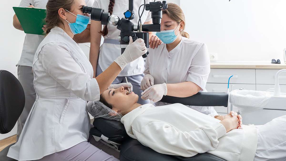 Patient reclined in dental chair receiving care from two masked clinicians in white coats.