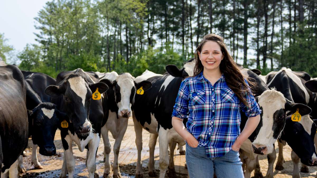 Mary stands with her hands on her hips in front of dairy cows with pine trees in background.