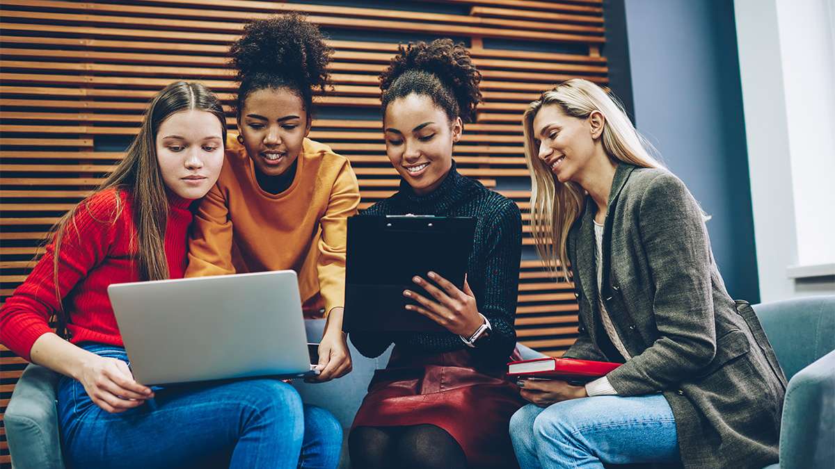 Four casually dressed young women of diverse backgrounds sitting together on a bench indoors, smiling and sharing a laptop.