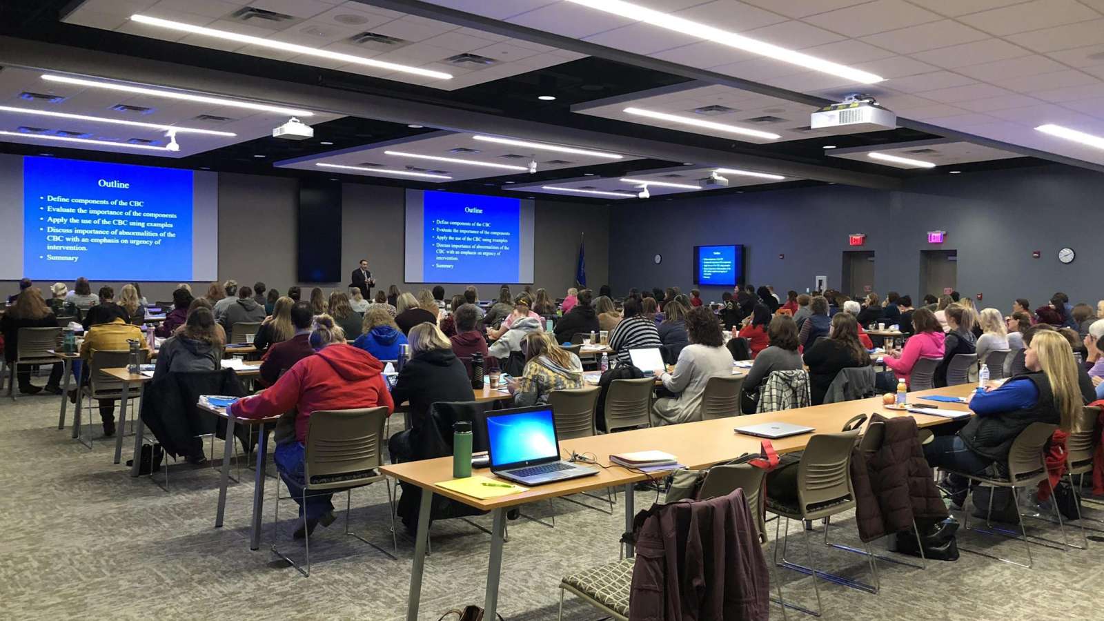 View from the back of the professional conference center with a speaker addressing conference attendees at the front of the room.