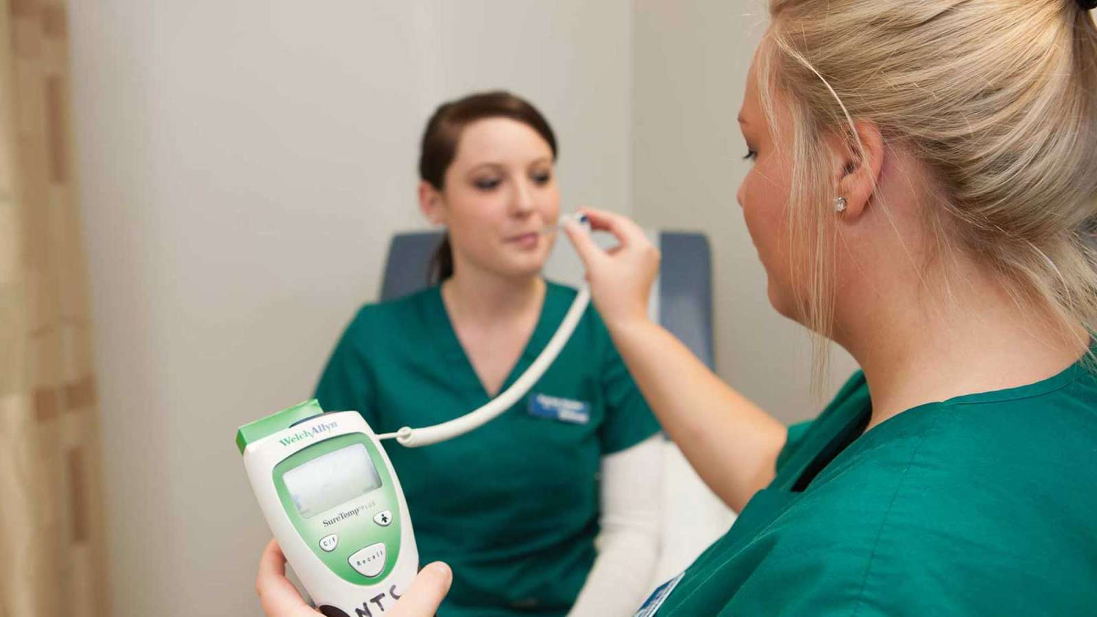 One student taking the temperature of another student in a clinic setting.