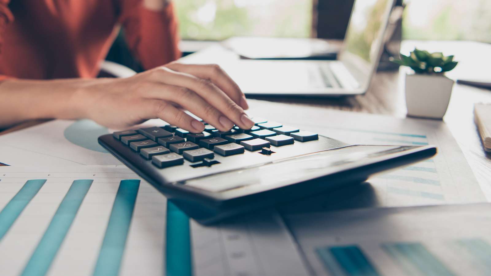 An accountant sits at a desk in front of a laptop calculating numbers with a calculator.