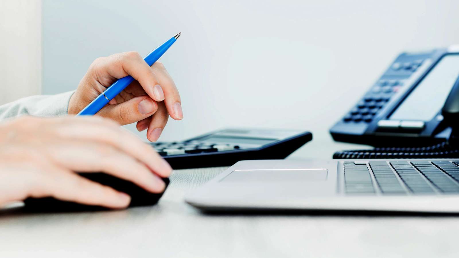 An accountant sits at a desk with a phone and a laptop while calculating numbers on a calculator.