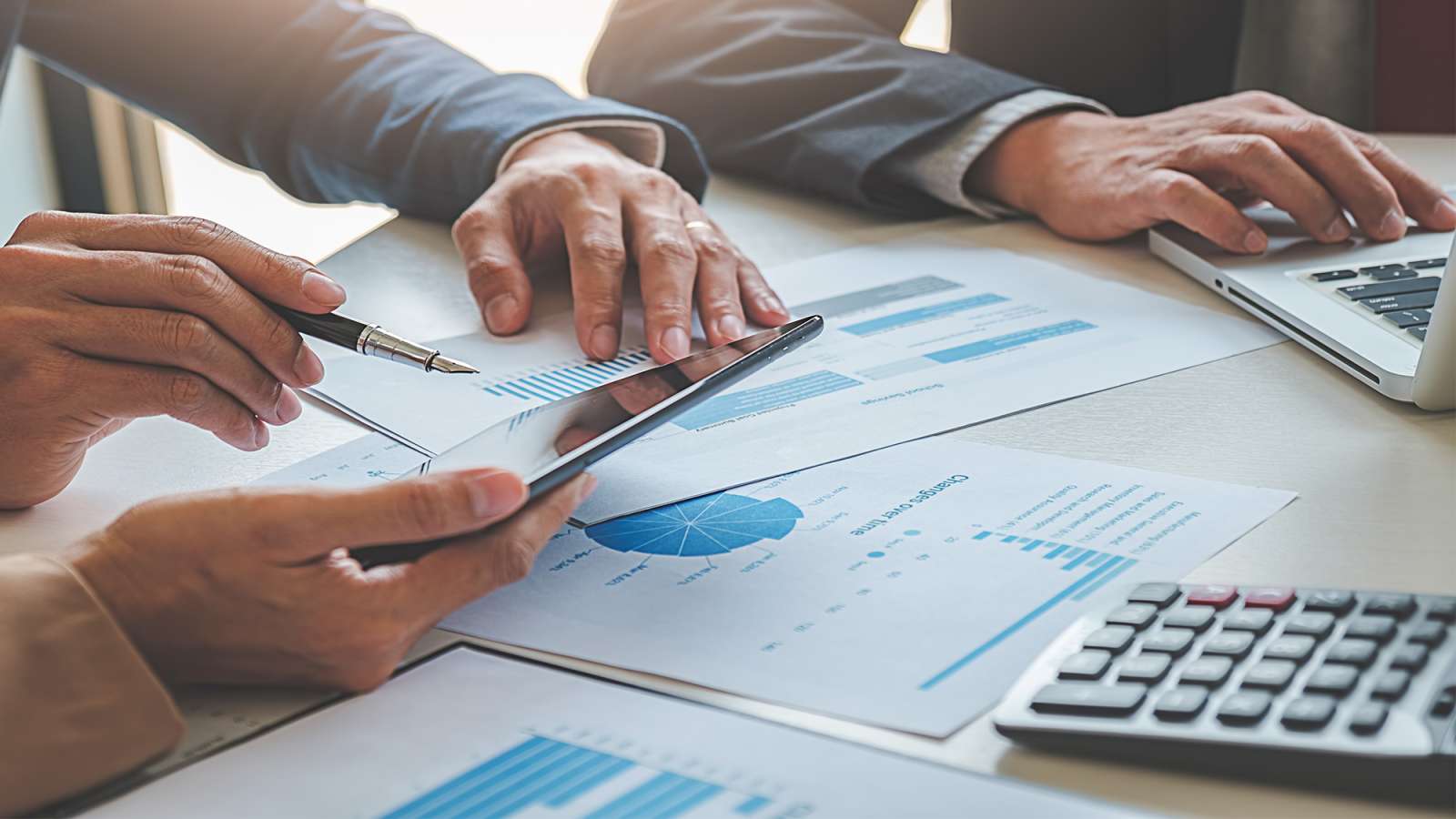 Several business persons sit at a table with spreadsheets, calculators, and a laptop