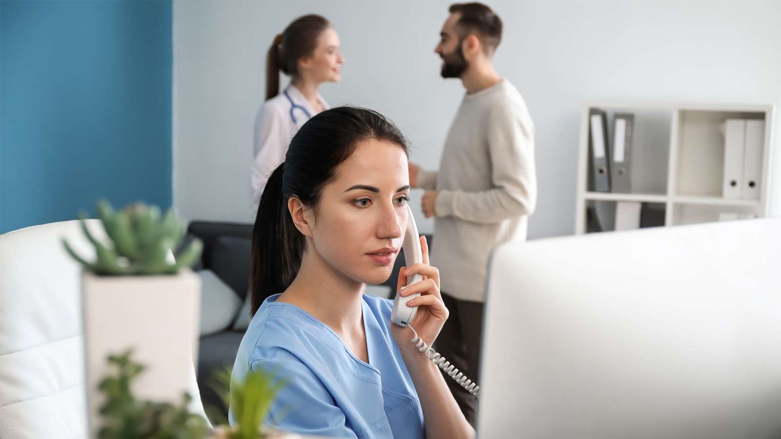 A doctor speaks with a patient in the background while a receptionist takes a phone call inside of a medical clinic.