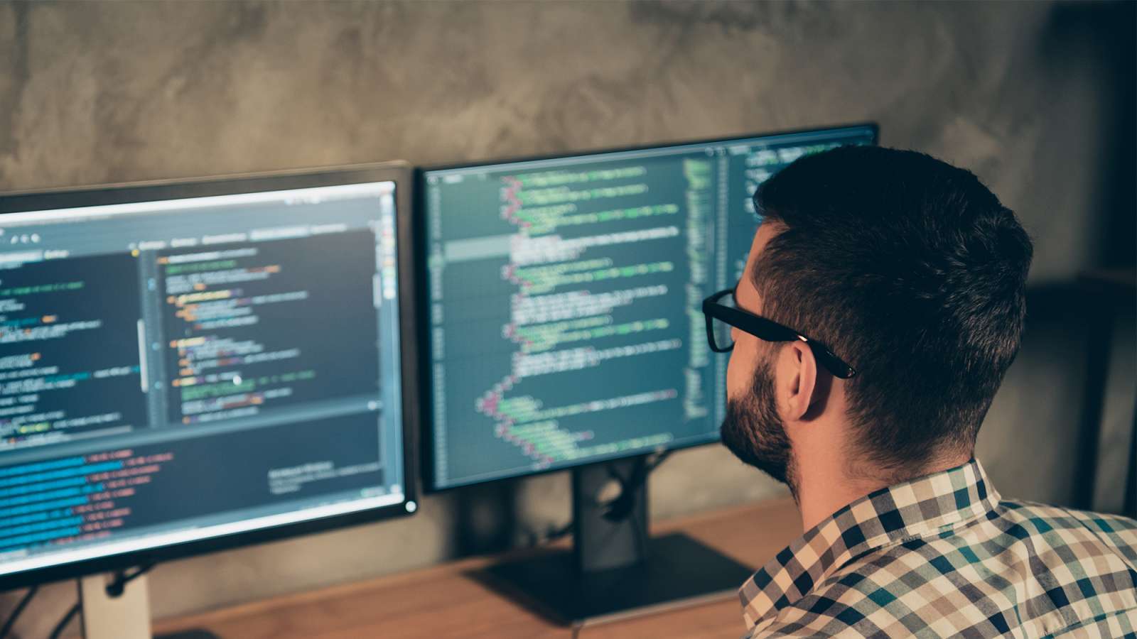 A man wearing a checkered shirt is sitting at a desk using a dual monitor setup to program an application.