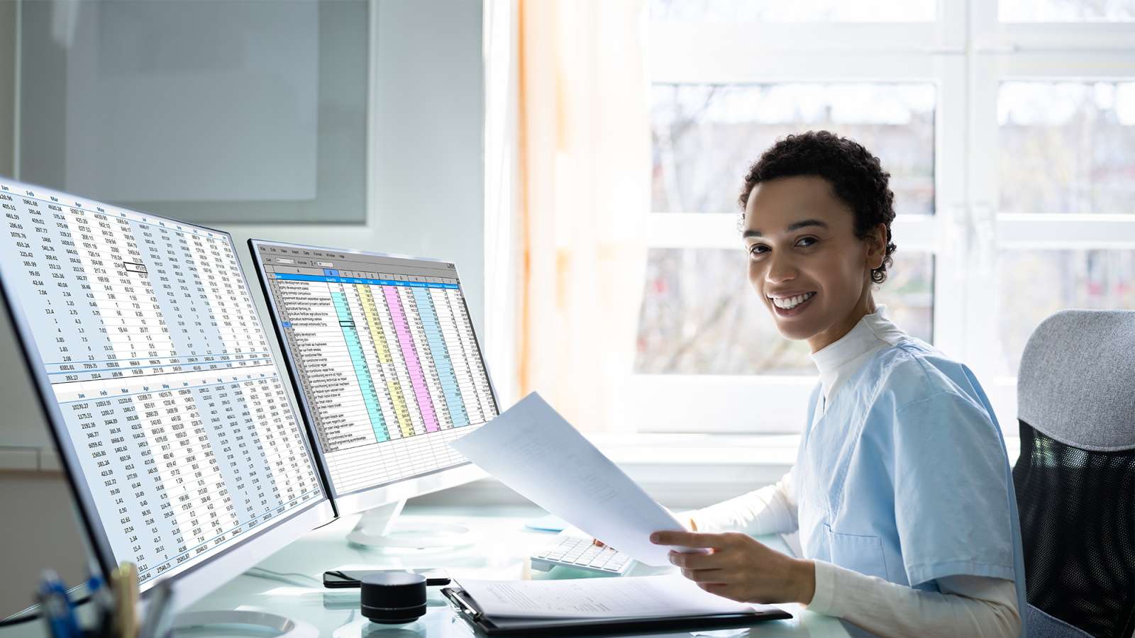 A medical coding specialist sits at a desk in front of a computer with two monitors and is holding documentation.