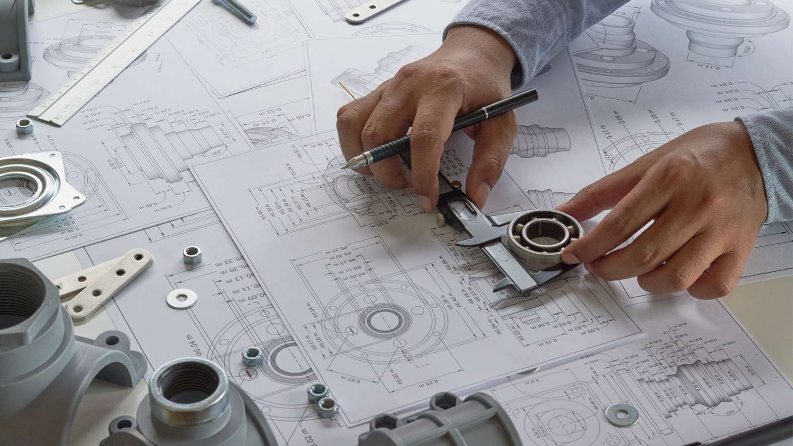 An Engineer technician is creating blueprint drawings of mechanical parts using parts set out on a desk for reference.
