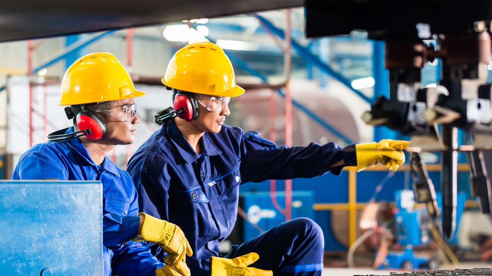Two people wearing safety helmets and blue jumpsuits are inspecting machinery inside of a factory.