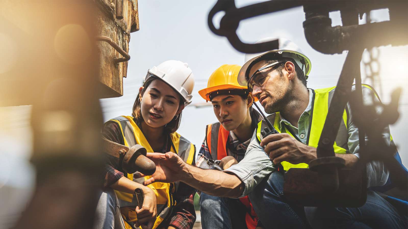 A team of three engineers wearing colored safety vests and hard hats inspect a broken train wheel.