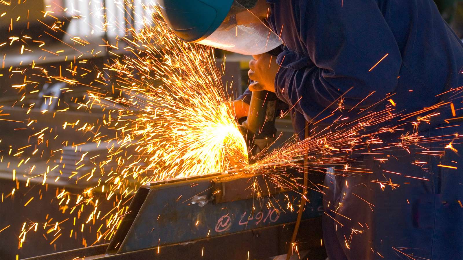 A Shielded Metal Arc Welder wearing a blue jumpsuit and face shield is cutting through metal inside of a factory.