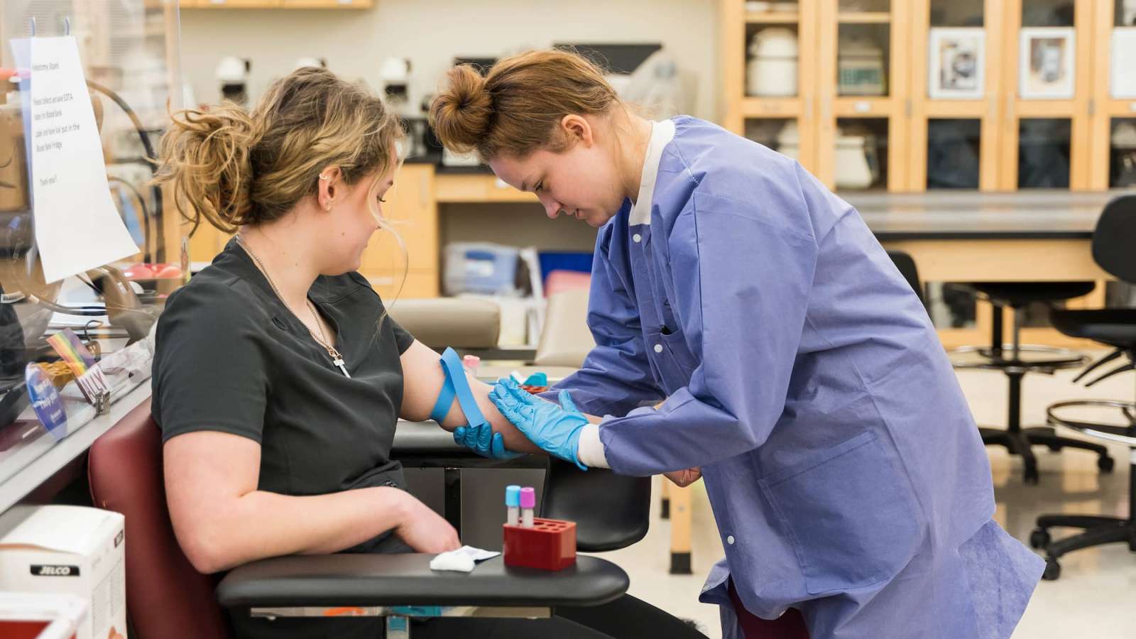 A view from the side of a Phlebotomy student placing their hand on a patient's forearm before drawing blood.