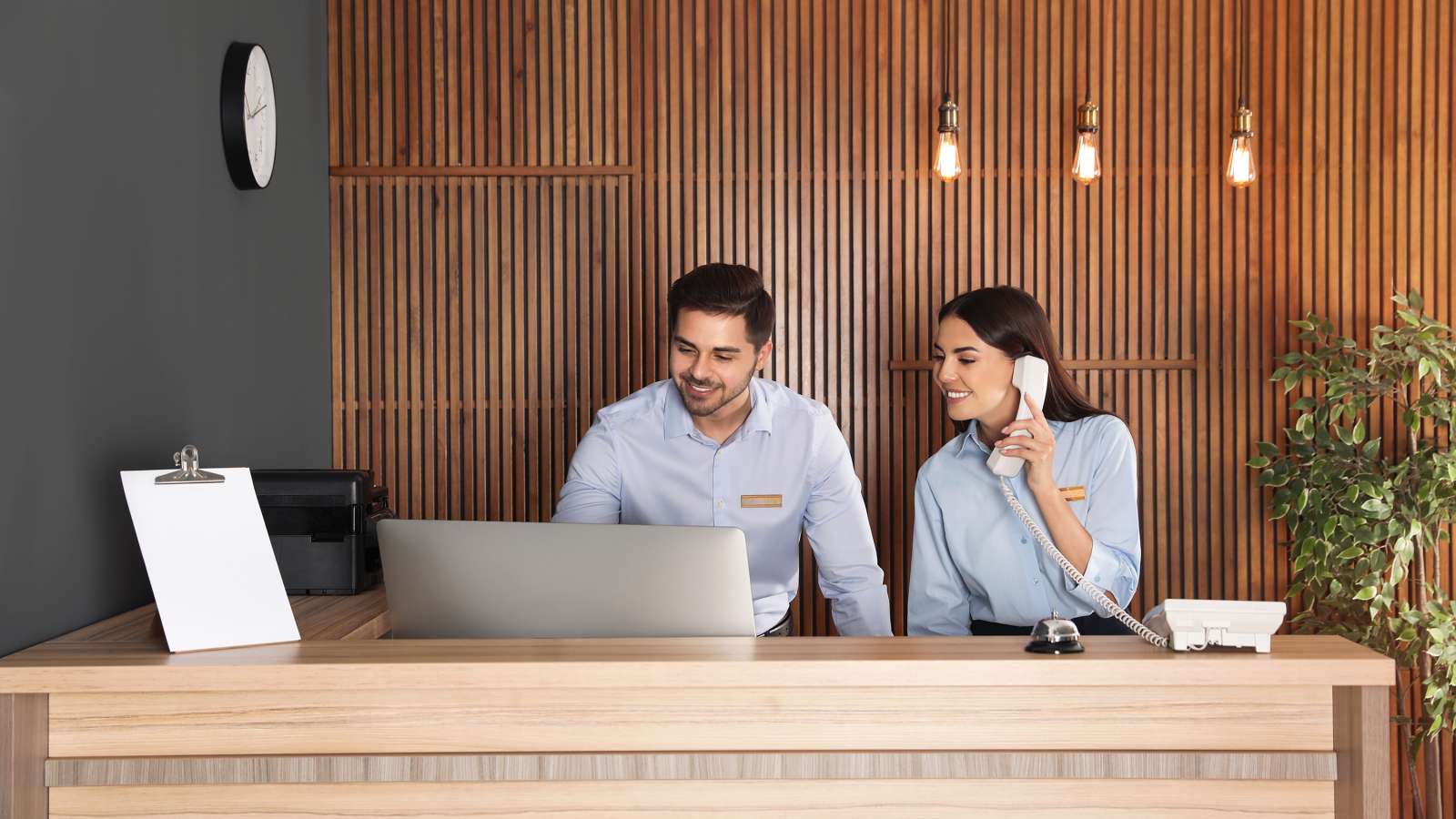 Two front desk employees working at a hotel. One employee is working on a computer and the other is talking on the phone.