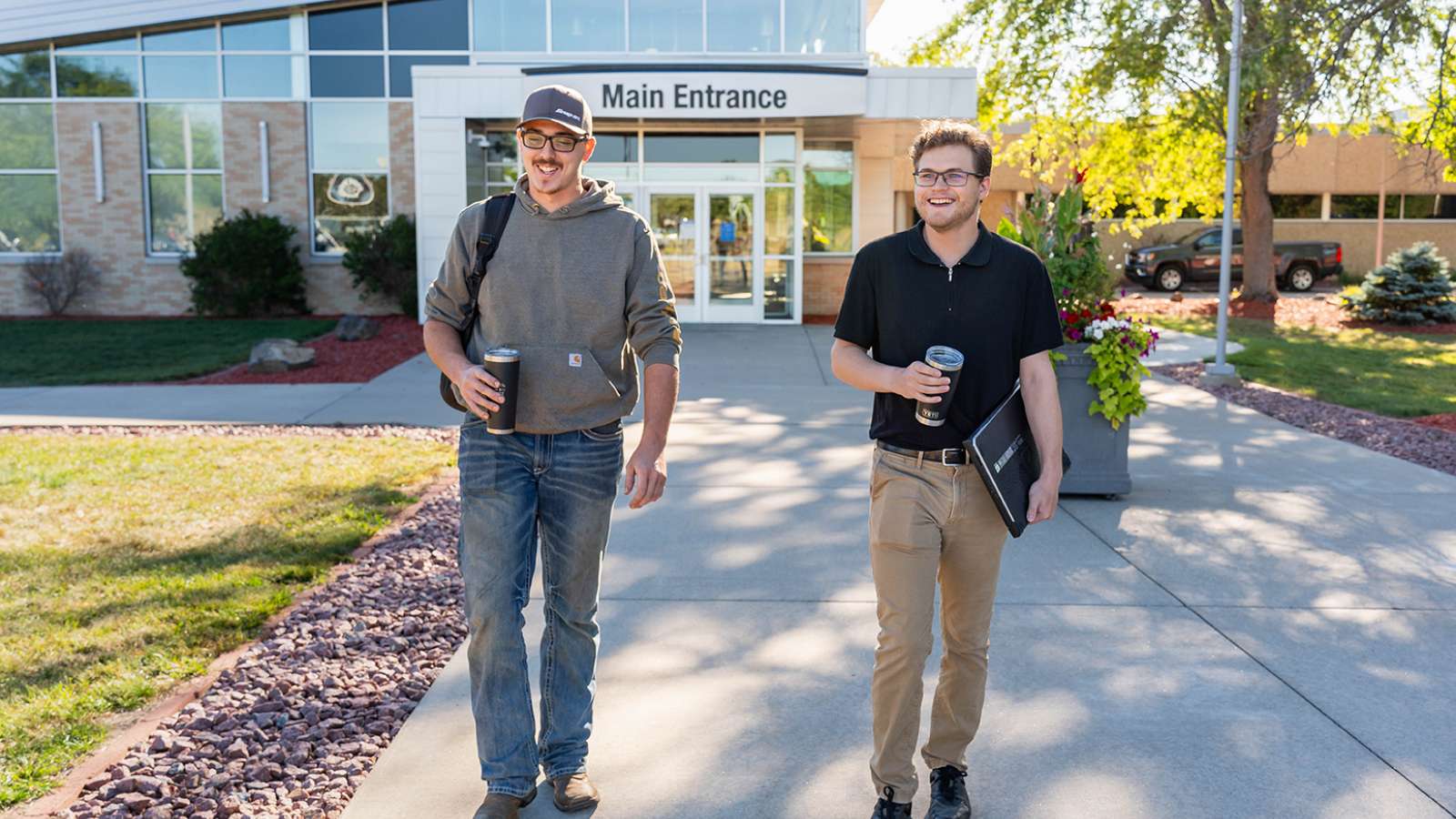 Two students sharing a laugh while carrying coffee mugs and school supplies as they walk outside the main entrance of the Wausau Campus.