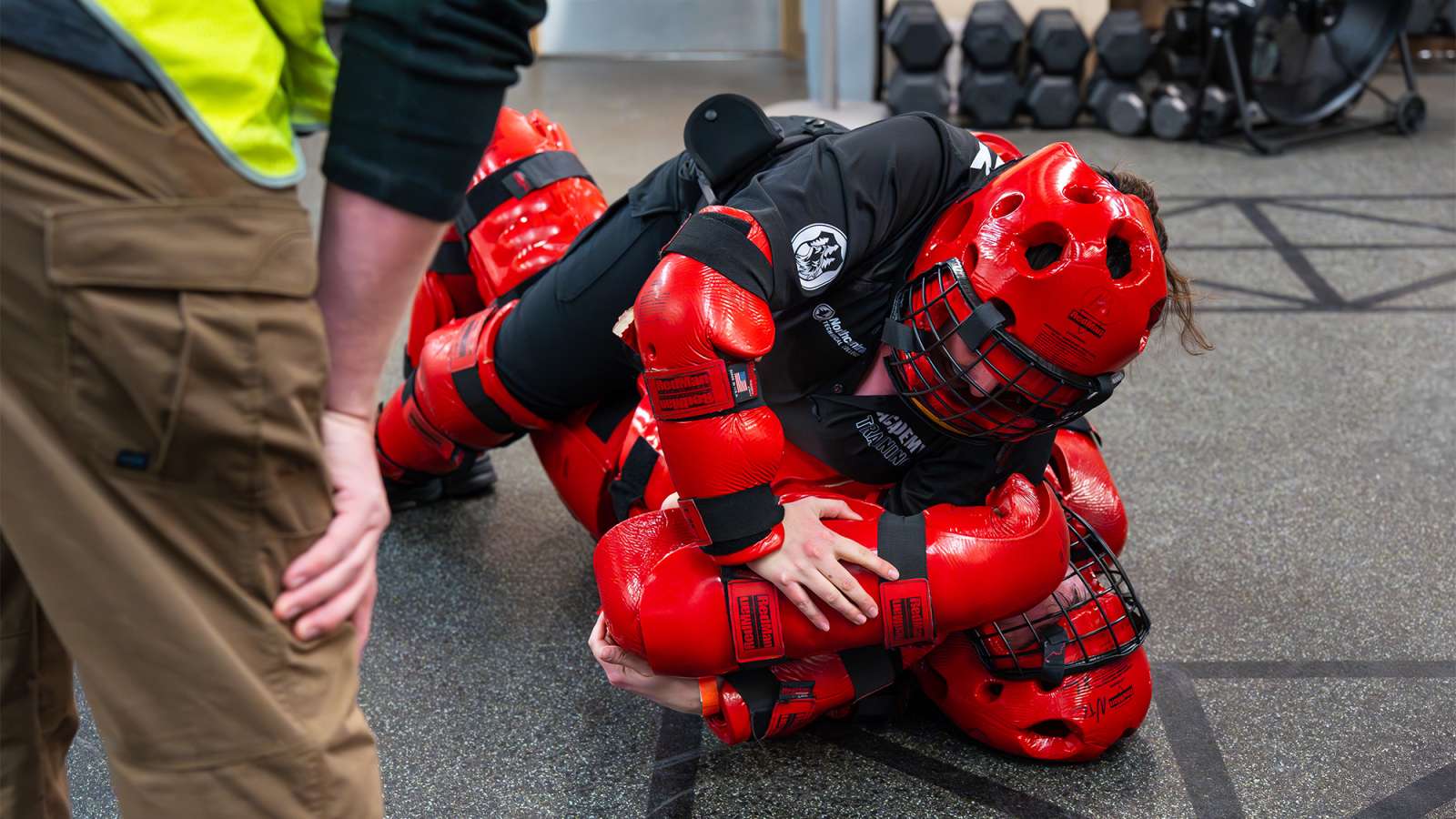 Two individuals in red padded gear practice ground defense techniques, supervised by an instructor in a training facility.