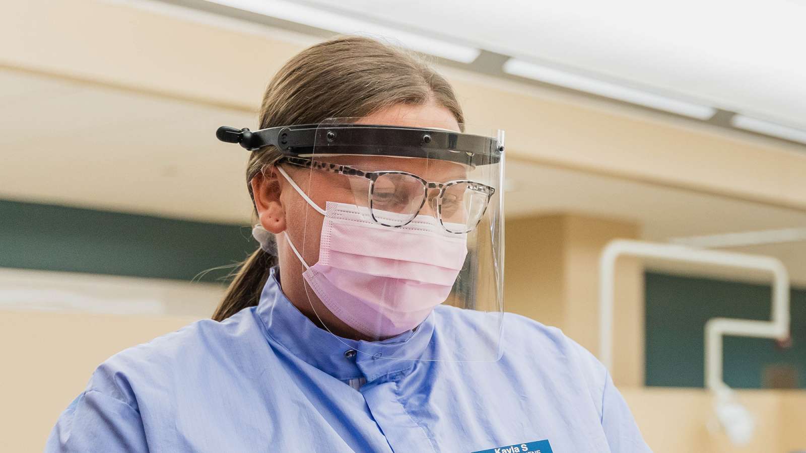 A healthcare professional wearing a face shield, mask, and protective gown in a clinical setting, focused on a dental task.