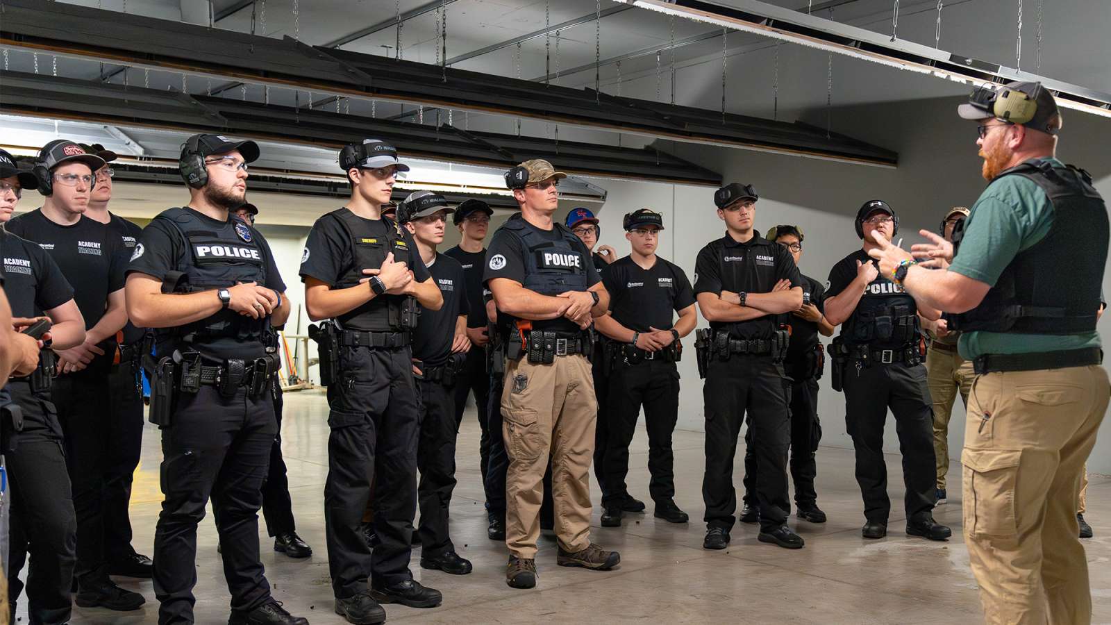 A group of police academy trainees in tactical gear attentively listen to an instructor during a training session indoors.
