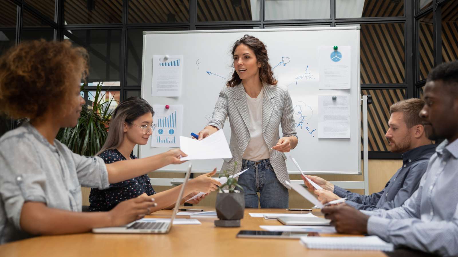 A woman in a blazer leads a meeting, handing out papers to a diverse team seated around a table with laptops. A whiteboard with charts is behind her.