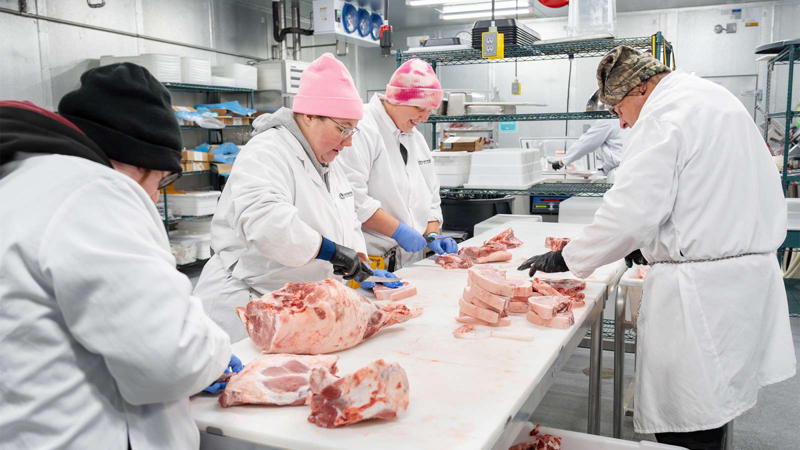 Four meat processing workers in white coats collaborate at a cutting table, processing various cuts of pork.