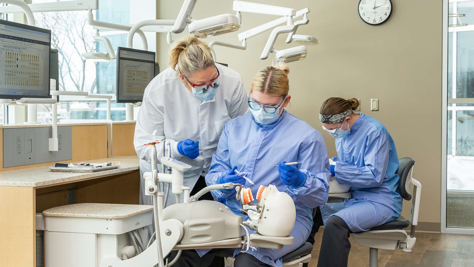 Dental instructor in a white coat guiding a student in blue scrubs practicing on a manikin, with another student nearby.