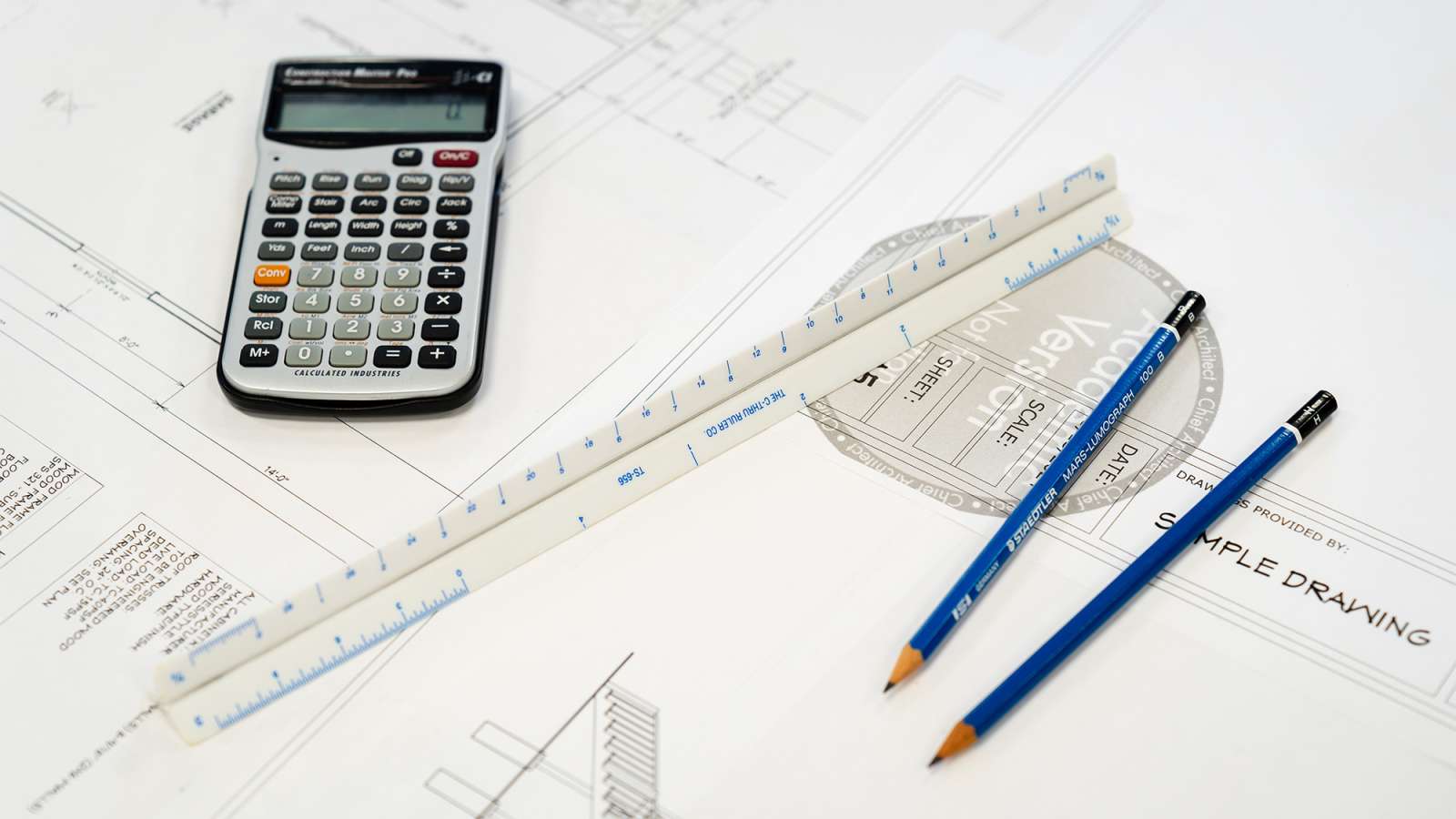 Engineering tools on architectural blueprint: calculator, rulers, pencils, and technical drawing document.