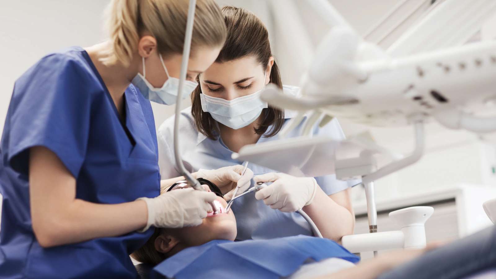Two dental professionals wearing scrubs, gloves, and face masks performing a dental procedure on a patient lying in a dentist’s chair, with dental instruments and overhead equipment visible.