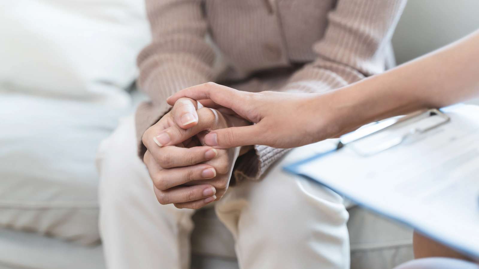 Close-up of a comforting gesture as one person gently holds another’s hands, with a clipboard in the foreground, symbolizing support, counseling, or advocacy in a compassionate setting.