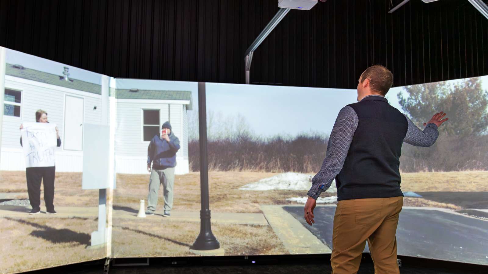 A man interacts with a large, curved screen in the Community Safety Simulation Center, participating in a virtual protest scenario with projected individuals holding signs and filming.