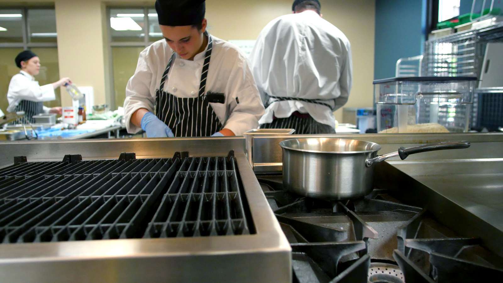 Culinary students in white uniforms and striped aprons work in a commercial kitchen, preparing food near a stove with a metal pot and grill in the foreground.