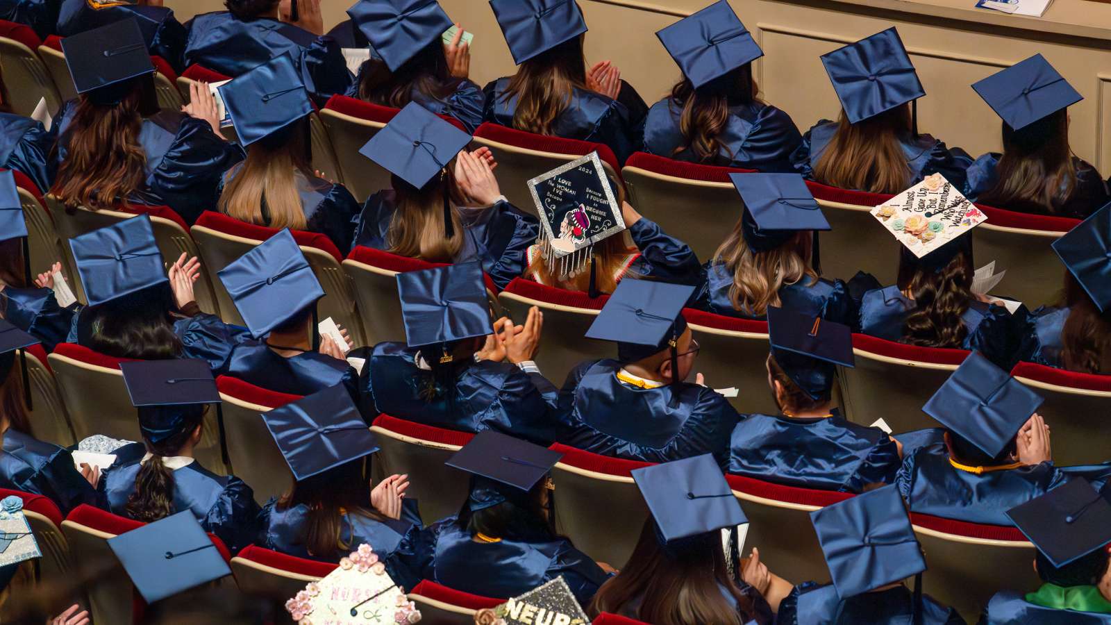 Students seated in their blue caps and gowns at a graduation ceremony at the Grand Theater, as seen from above.