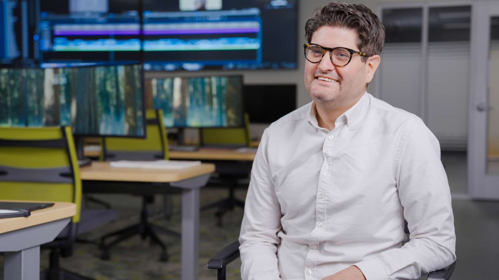 Jordan smiles, while seated in a video production room, with multiple computer monitors behind him displaying video editing software and nature scenes.