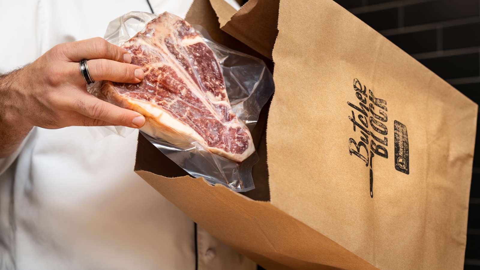 Butcher Block staff’s hand placing a vacuum-sealed T-bone steak into a brown paper bag displaying "The Butcher's Block" logo.