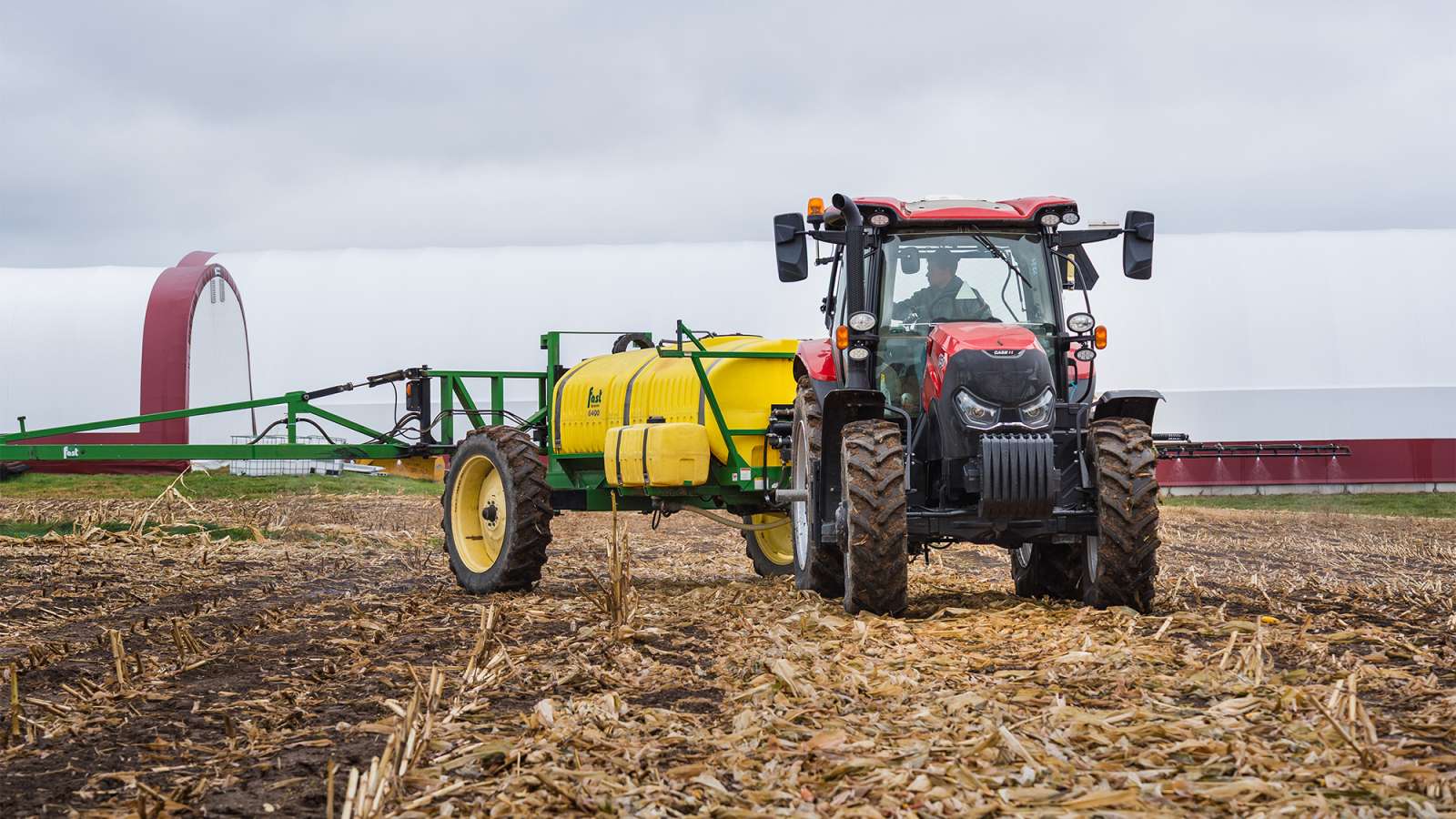 Red tractor in harvested cornfield towing a yellow sprayer with green booms under an overcast sky.