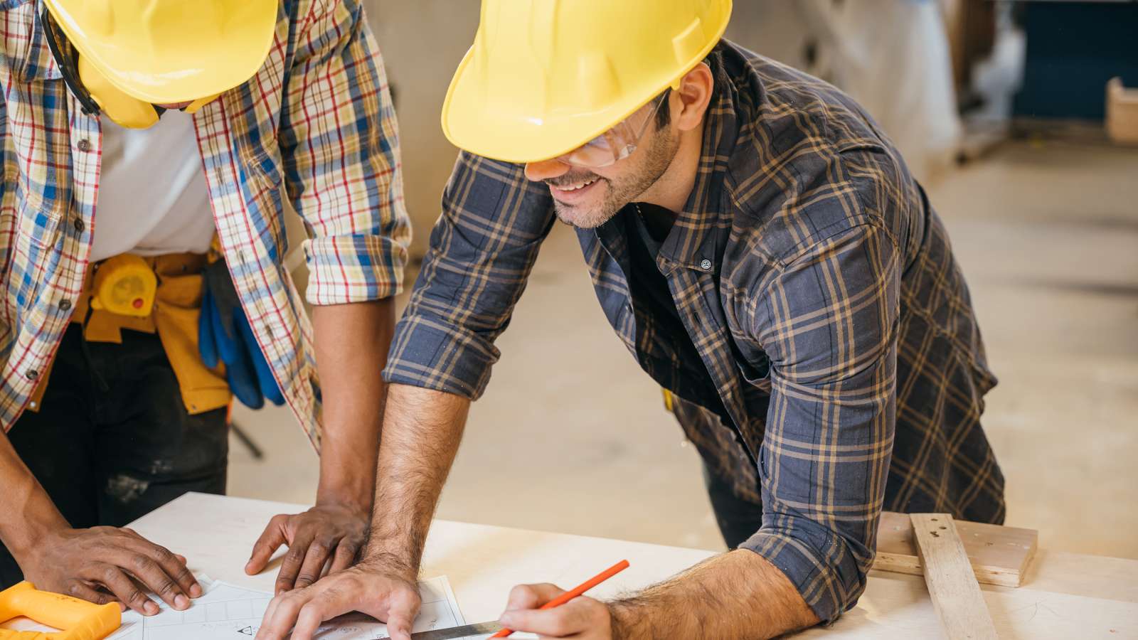 Two carpenters wearing yellow hard hats and plaid shirts lean over blueprints on a workshop table, pointing and writing amid lumber and tools.