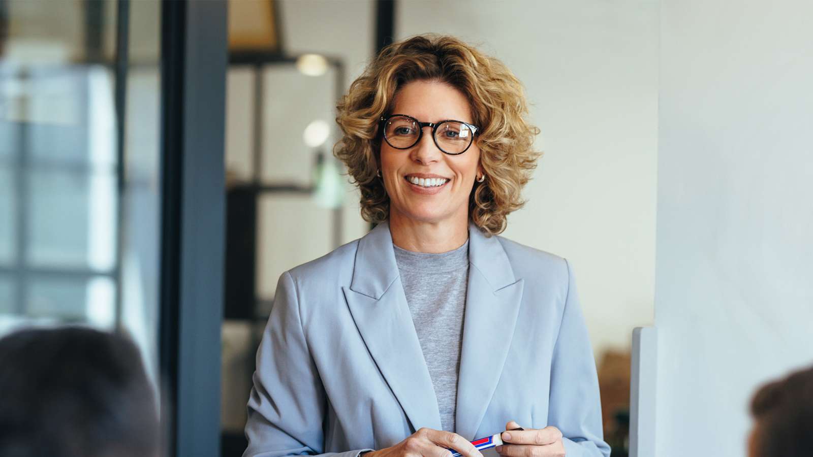 Smiling middle-aged woman with curly blonde hair and glasses in a light gray blazer, holding a pen, speaking with colleagues.